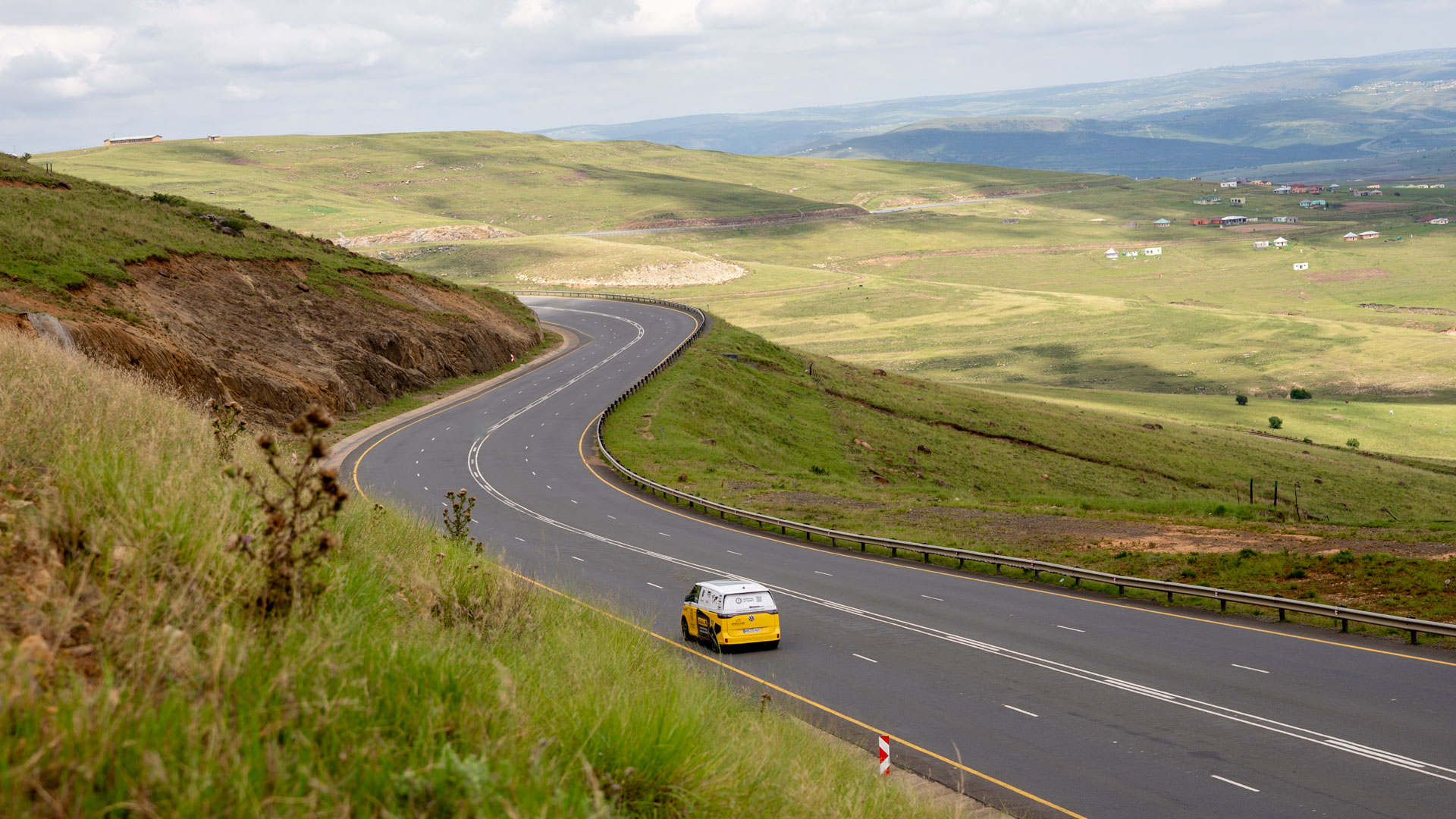 A yellow and white VW ID bus drives along a country road through green countryside.