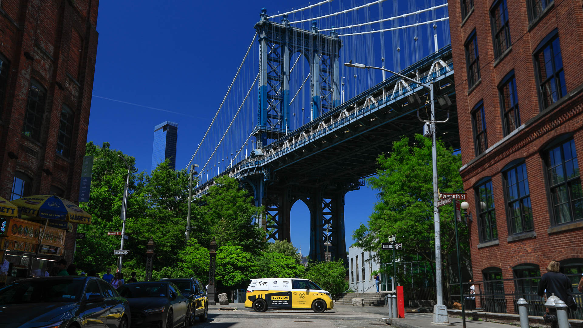 ellow and white Volkswagen electric microbus parked on a street in DUMBO, Brooklyn, beneath the Manhattan Bridge, with brick buildings, trees, and city traffic under a clear blue sky.