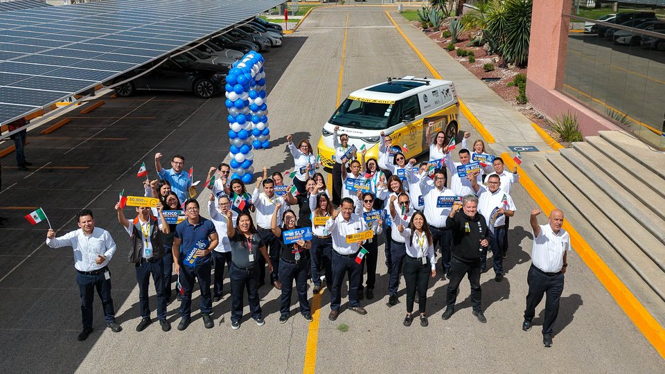Group of employees with handsigns and Mexican flags gathered outdoors on a lawn, posing together near a branded Volkswagen electric microbus in a parking lot.