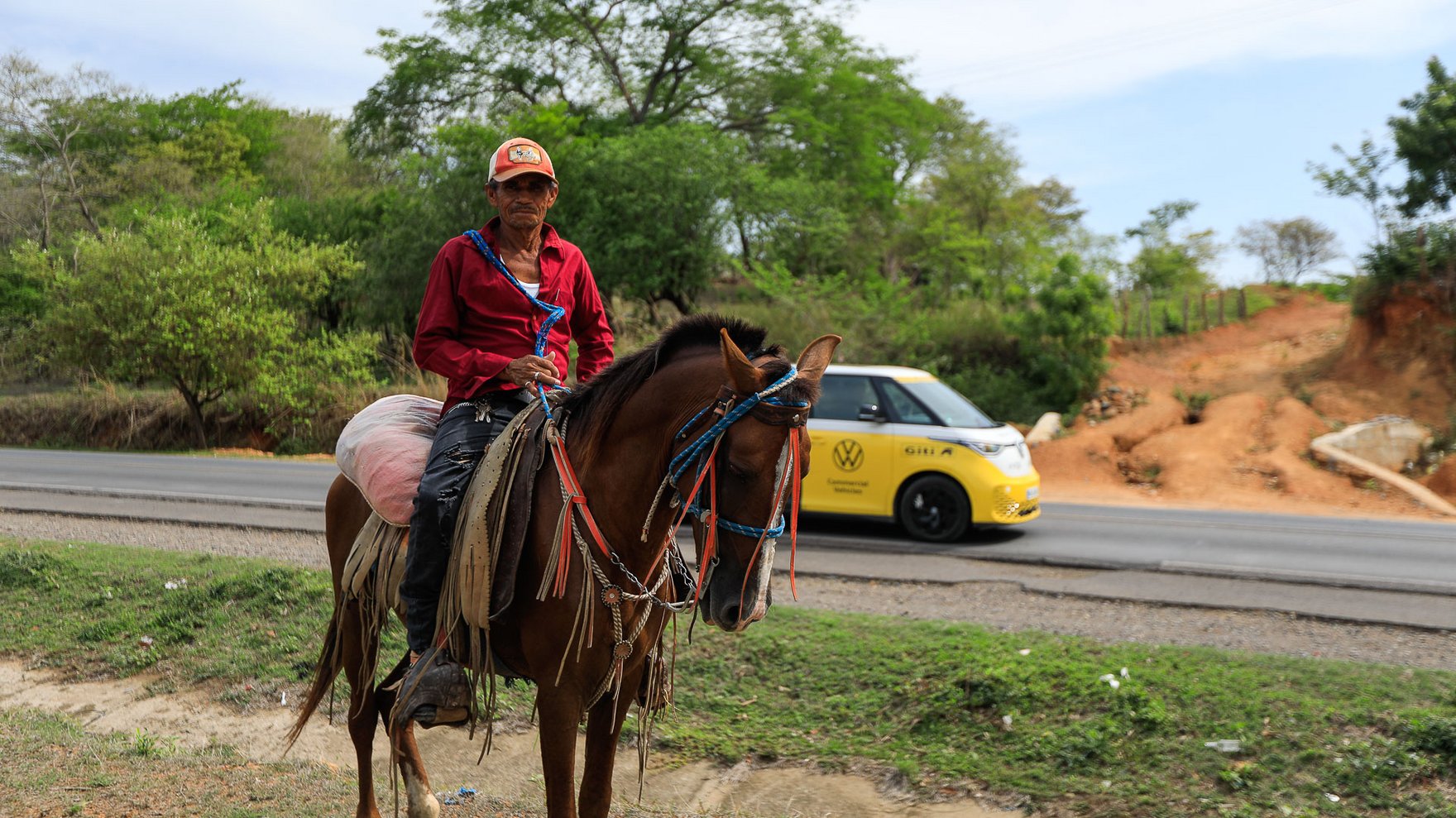 Man wearing a red baseball hat, a red longsleeve shirt and black pants sitting on a brown horse, next to a road on which a yellow van is driving. green trees and brown dirt road in the background