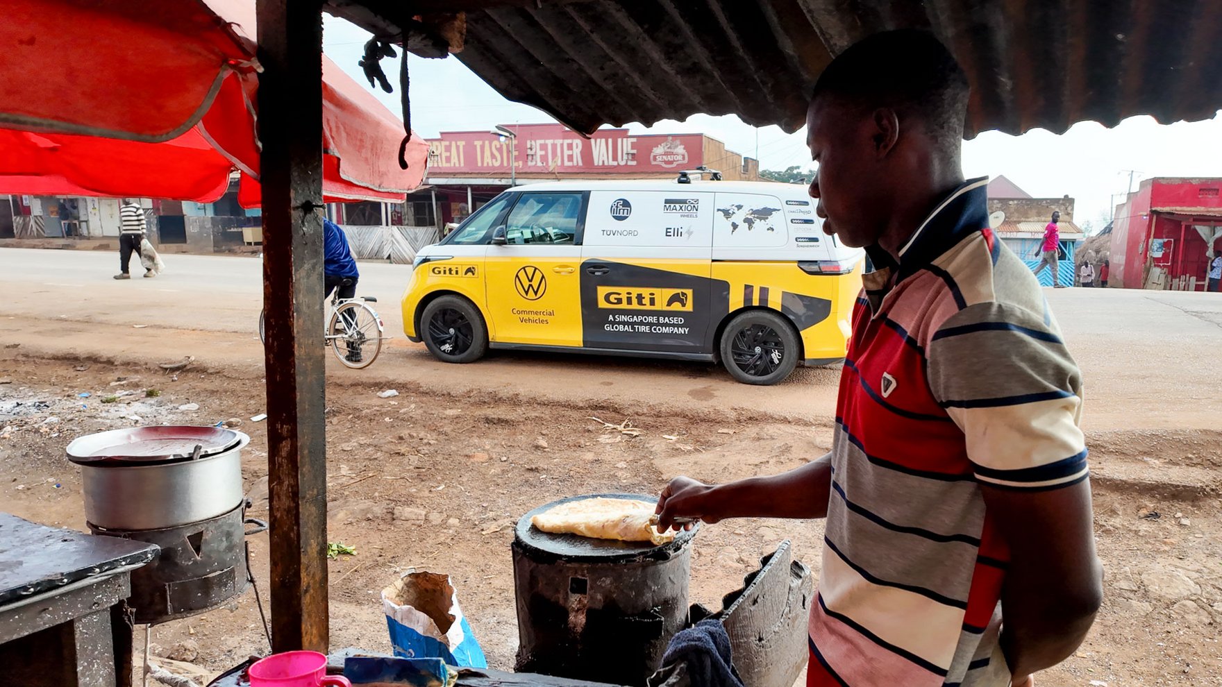 A black man is vooking in a hut, in the background a yellow and white VW ID bus is passing on the road 