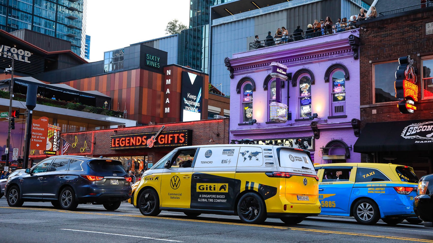Yellow and white Volkswagen electric microbus driving through a lively city street in Nashville, with neon signs, storefronts, and traffic, including a taxi, under evening light.