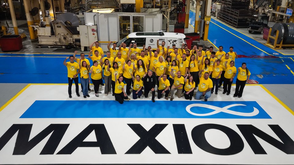 Large group of factory employees wearing yellow shirts pose on a production floor in front of industrial machinery and a branded Volkswagen electric microbus, with “MAXION” text visible on the ground.