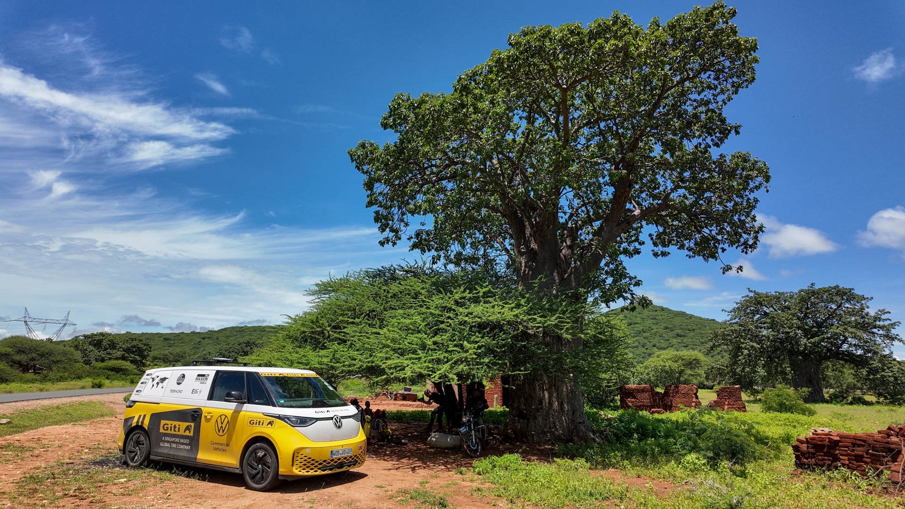 A yellow and white VW ID bus is parked next to a very large green tree, blue sky in the background