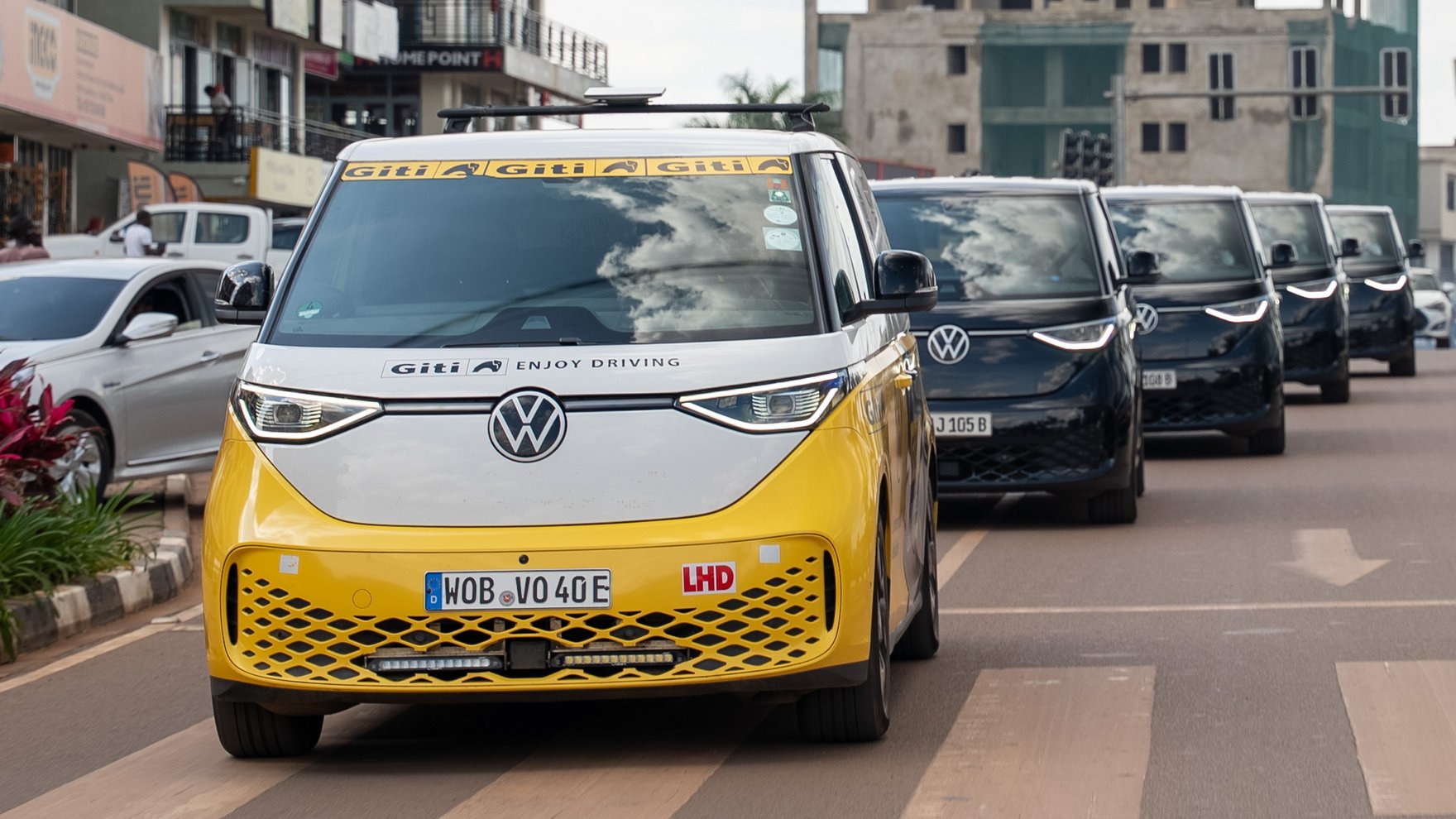 A yellow and white VW ID bus is driving in a city, followed by 4 black VW ID bus vehicles