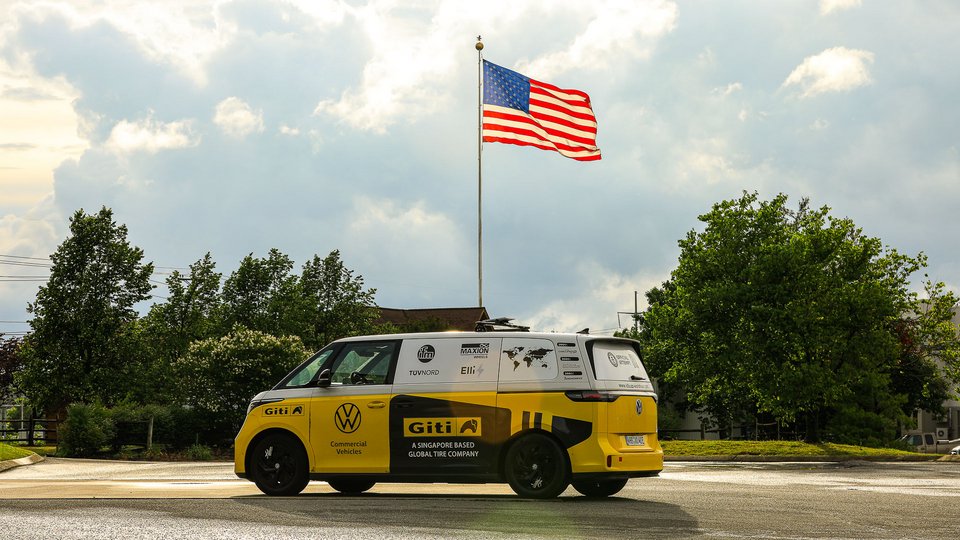 Yellow and white Volkswagen electric microbus parked on a roadside beneath a large American flag on a tall pole, with green trees and a cloudy sky in the background.