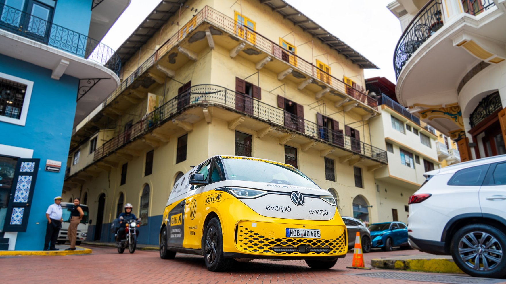 yellow volkswagen van driving through narrow red brick street running through 3 story stone buildings in blue, yellow and white. some cars parked on the street, a motorcycle is driving behind the van and two pedestrians are looking on