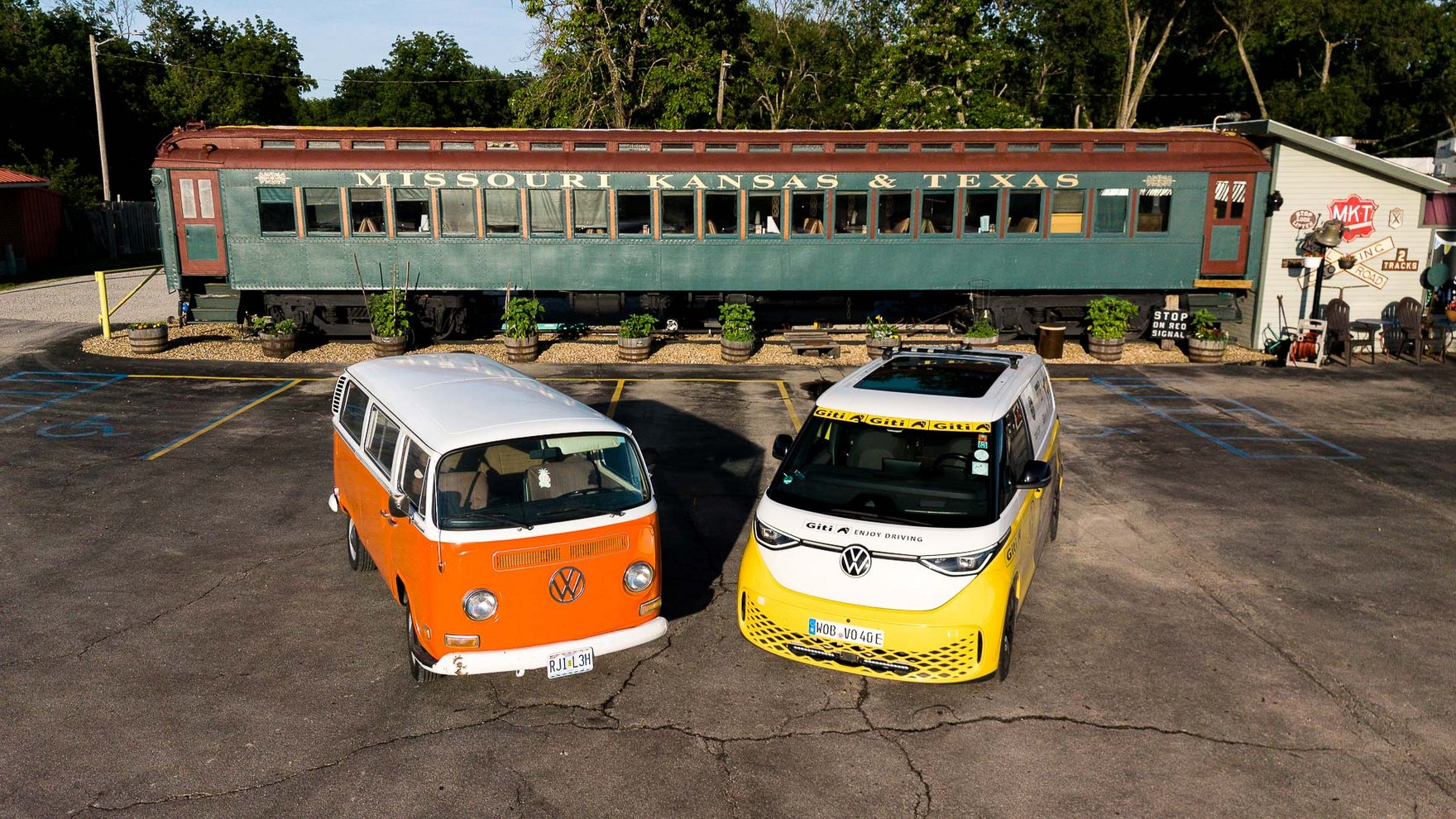 Vintage orange and white Volkswagen van parked beside a modern yellow and white electric Volkswagen microbus in front of a historic “Missouri Kansas & Texas” railcar, with trees and a parking area in the background.