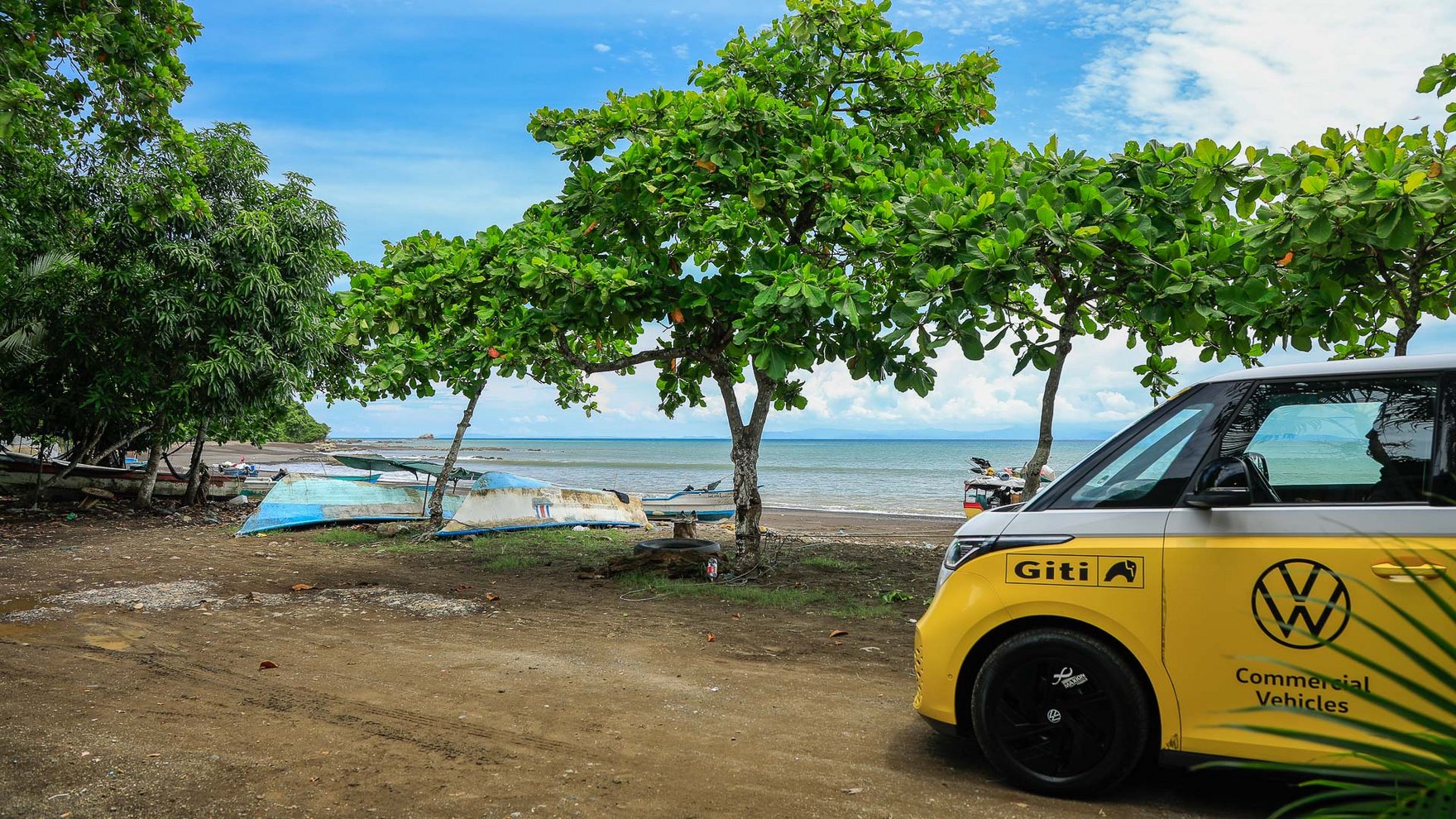 yellow van parked on a sandy road next to a beach, blue and white boats sitting on the sand, green trees lining the beach and the turquoise ocean in the back