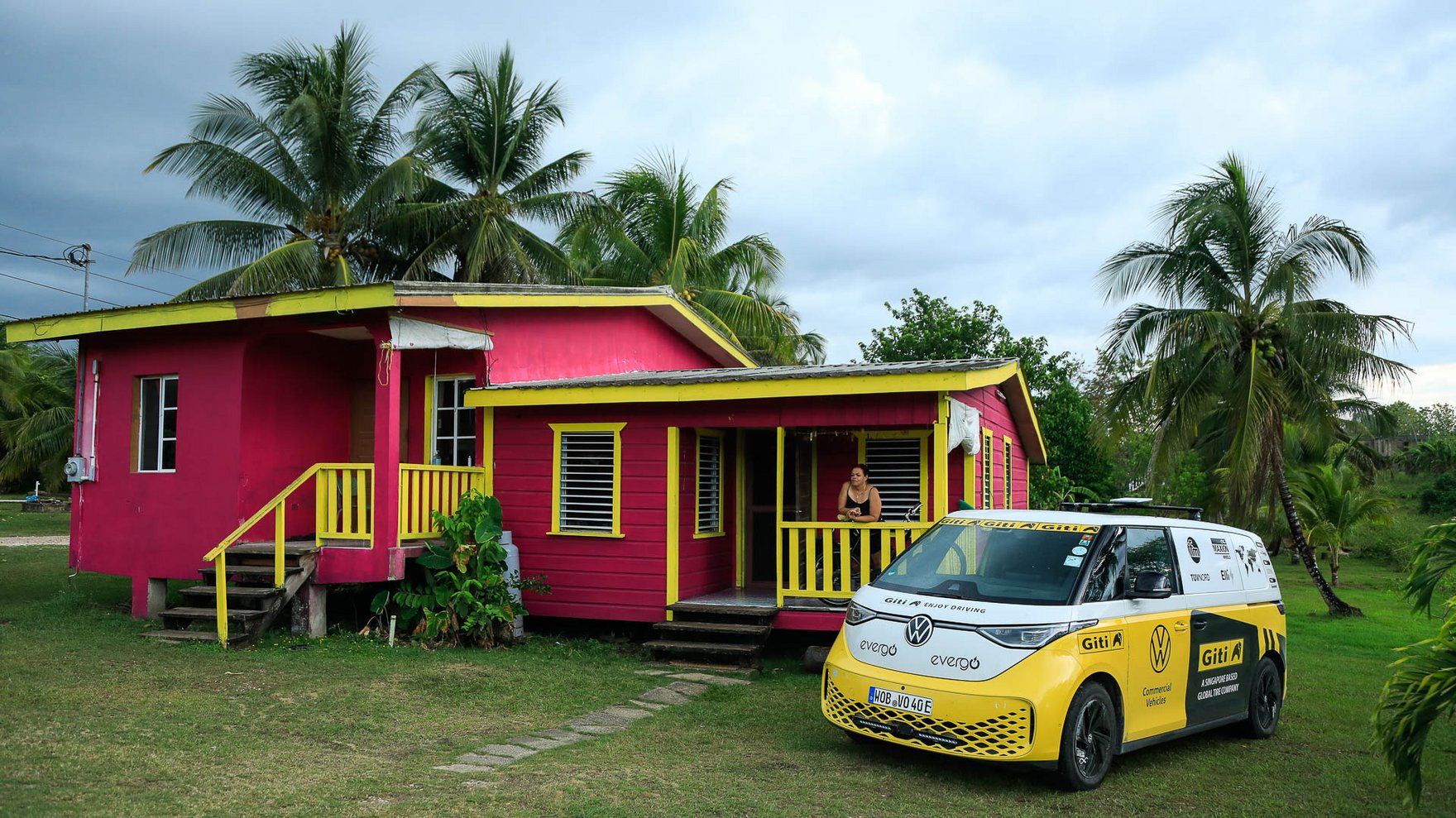 yellow Volkswagen ID Buzz parked on green grass next to a wood house in pink and yellow, palm trees in the background