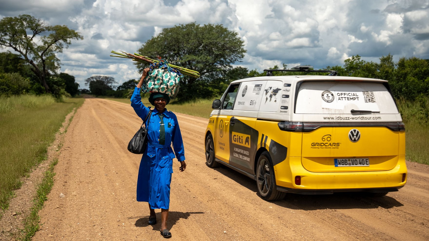 A yellow and white VW ID bus is driving on a dirt road, a black woman wearing a blue dress and carrying a bundle on her head is walking next to it