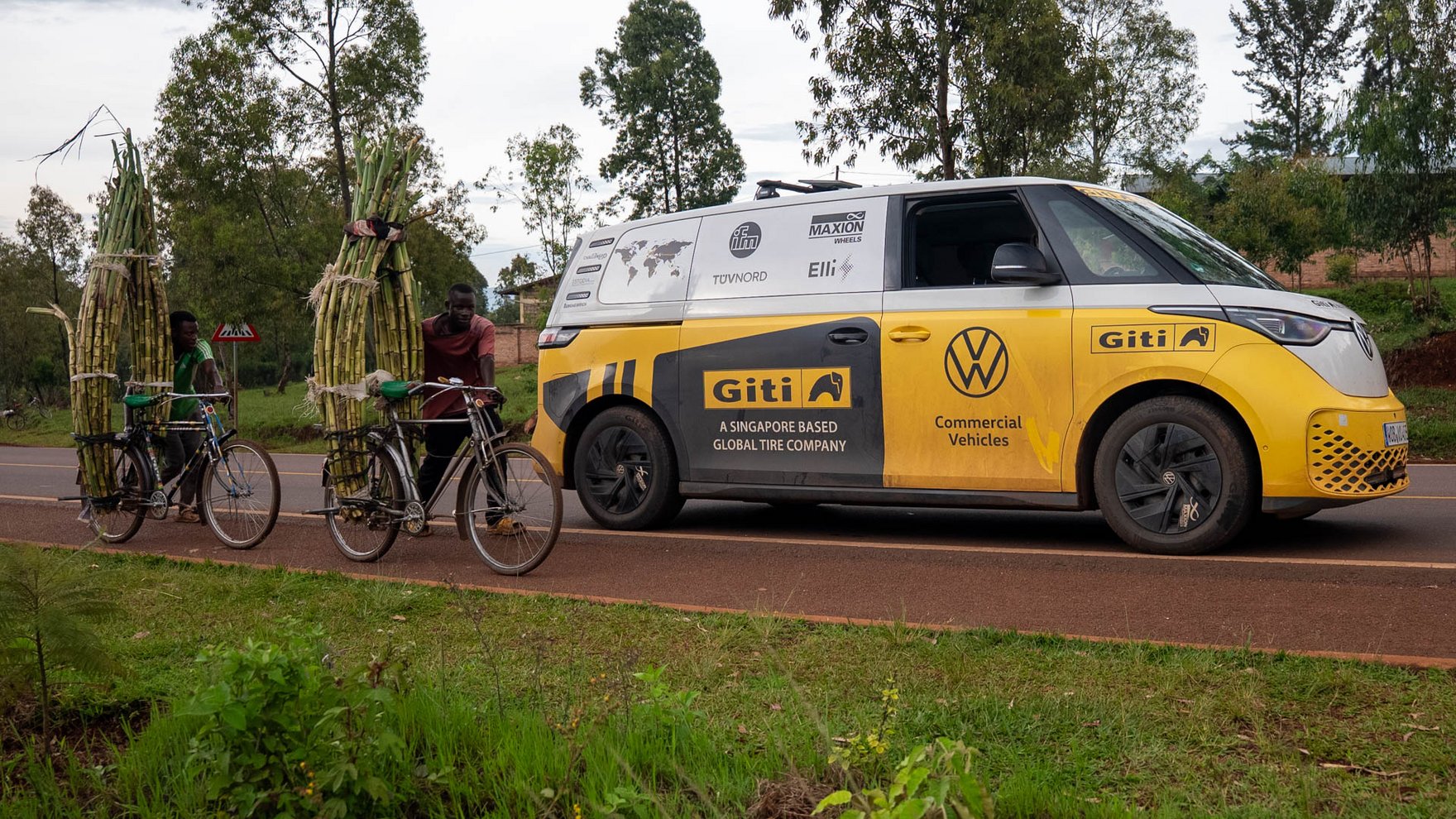 A yellow and white VW ID bus is parked on a country road, next to it two men are pushing their bicycles transporting green bamboo sticks