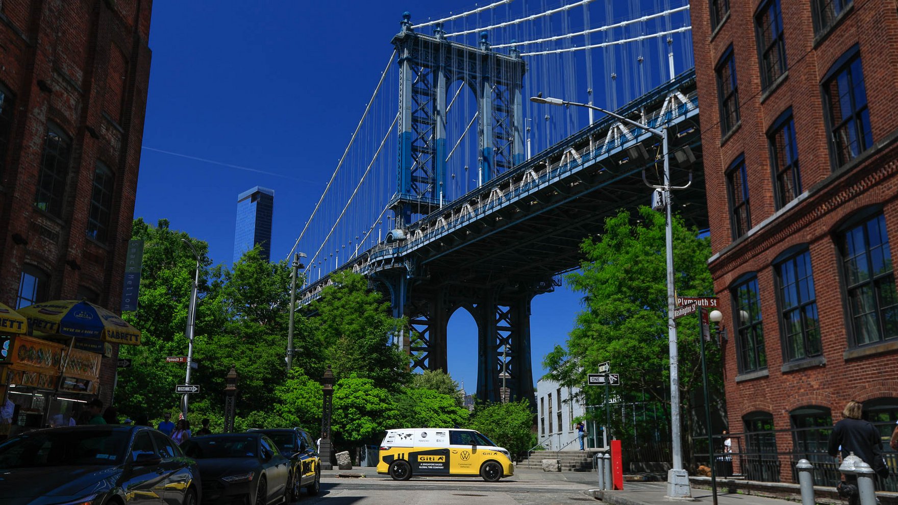 ellow and white Volkswagen electric microbus parked on a street in DUMBO, Brooklyn, beneath the Manhattan Bridge, with brick buildings, trees, and city traffic under a clear blue sky.