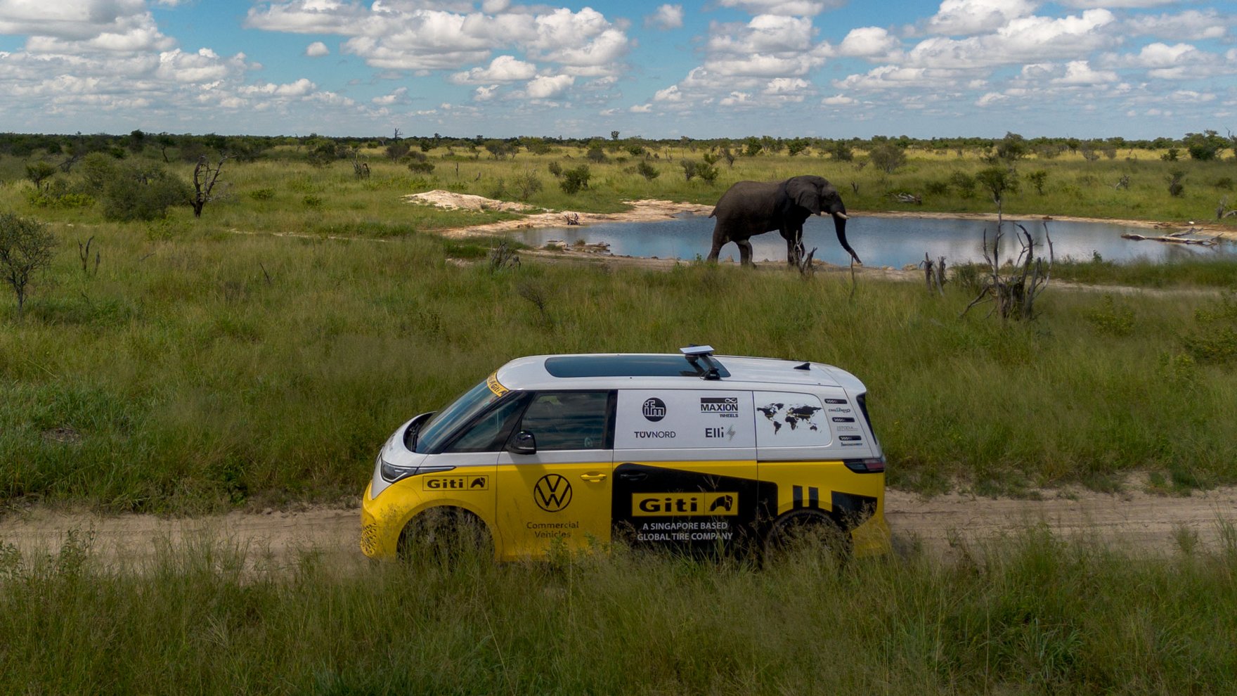A yellow and white VW ID bus is parked in an open green field, an elephant is standing next to a waterhole