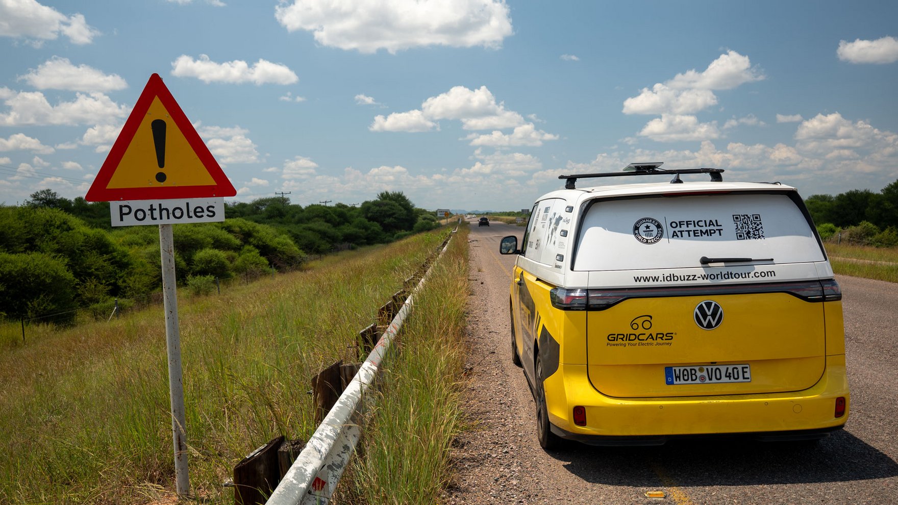 A yellow and white VW ID bus is driving on a country road, a triangular shaed yellow and red road sign warns drivers of potholes