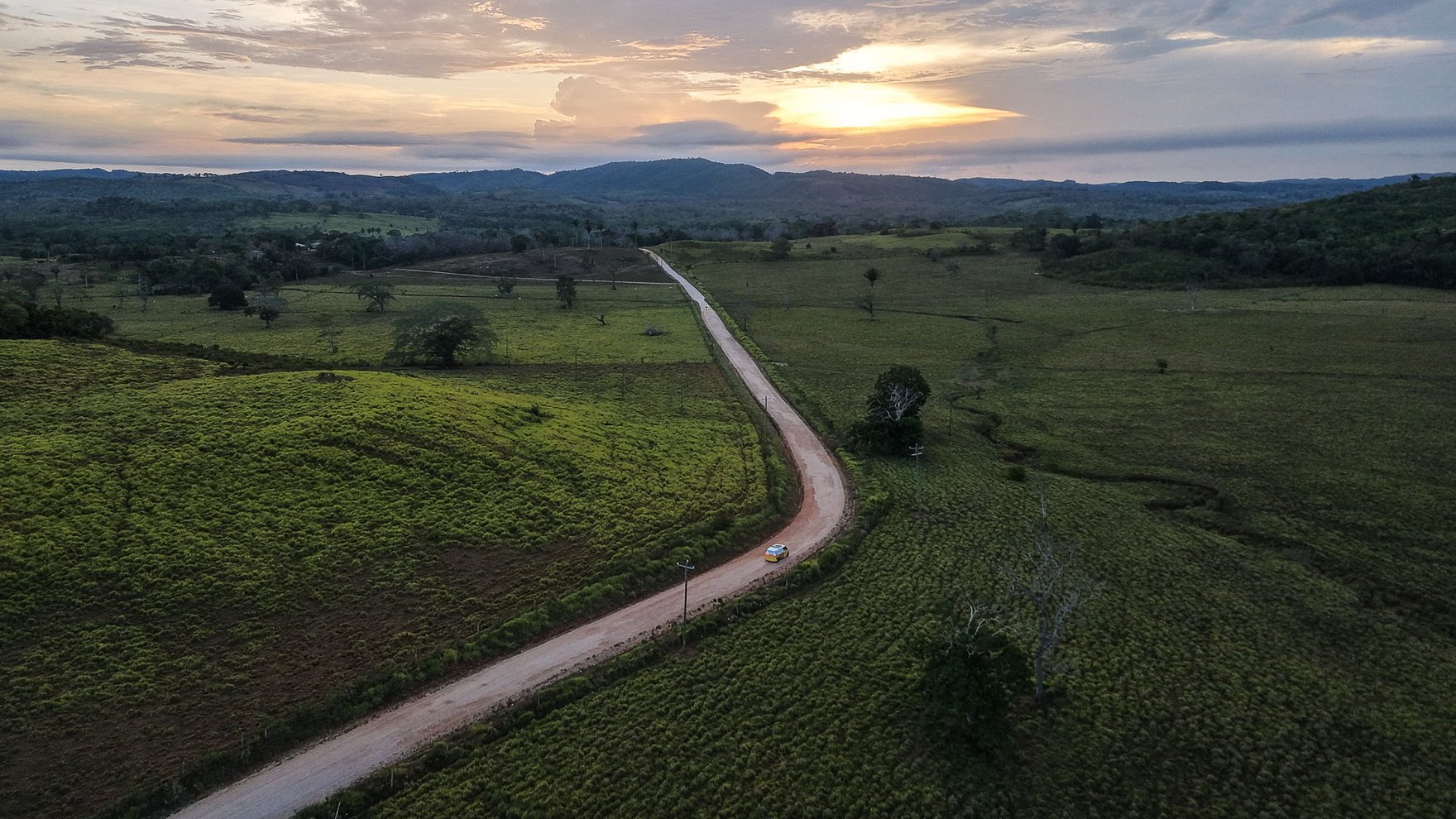 view from an elevated viewing point into the distance, a road cutting through green fields, the sun is setting behind clouds on the blue and yellow horizon