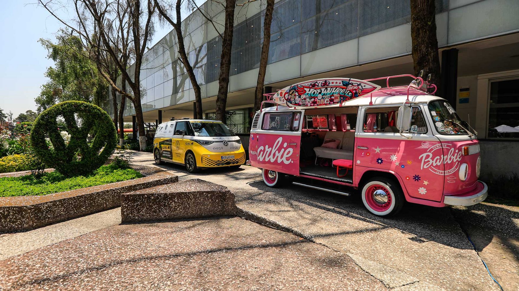 decorated pink Volkswagen van with Barbie logo, Volkswagen logo carved in a bush and yellow van parked in the background